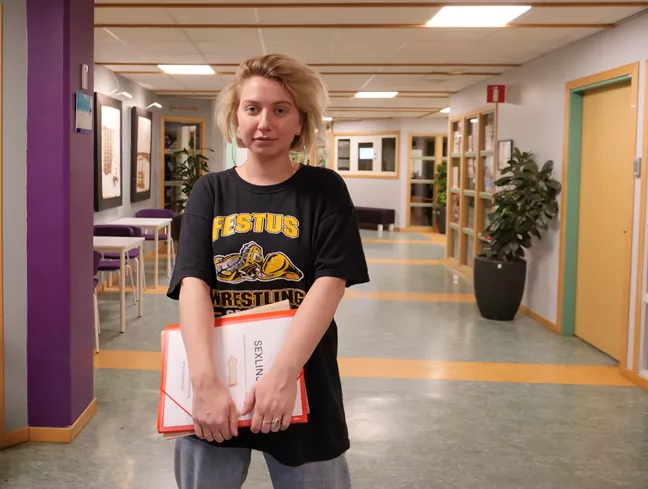 Linda står i skolkorridoren och tittar in i kameran. 